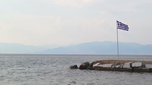 Greek flag in the wind, rocky shore and mainland in far distance, useful for current event video backgrounds related to Greek monetary crisis and disputes with European Union