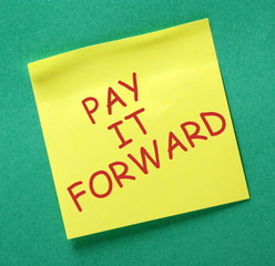 The phrase Pay It Forward in red text on a yellow sticky note posted on a green notice board
