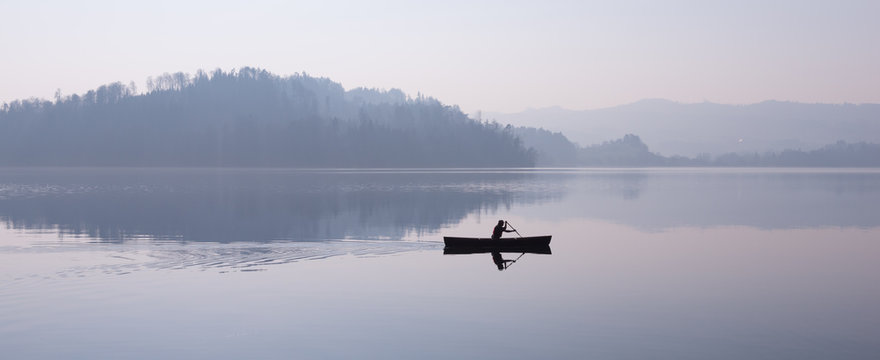 Man floating in a boat. Fog over the lake, mountains reflected in the water