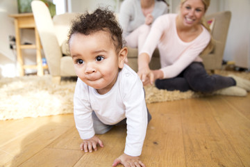 Female couple watch proudly as their baby son crawls along the floor - obrazy, fototapety, plakaty