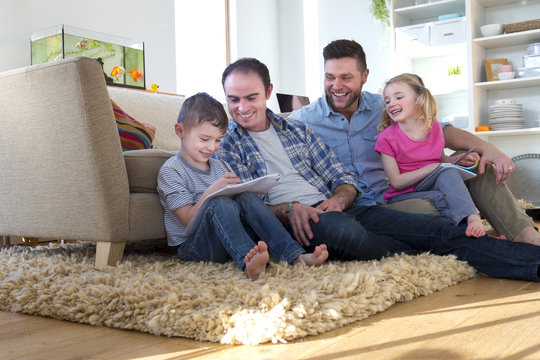 Same sex male couple sitting on the floor of their front room with their children, helping them do their homework