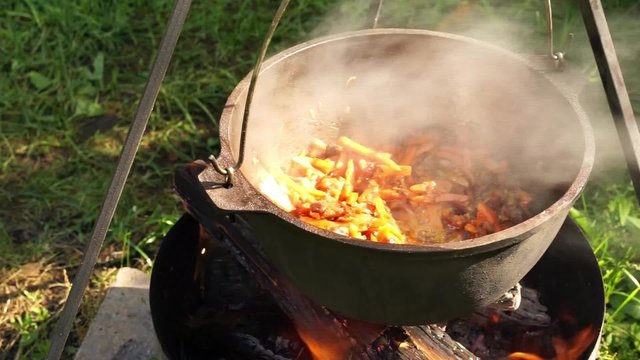 close up view of cooking meat, onion and carrot in a metal pan in a campfire