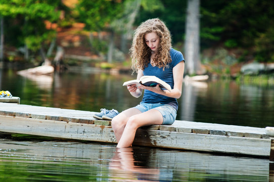 Teen Girl Sitting On A Dock Reading A Book