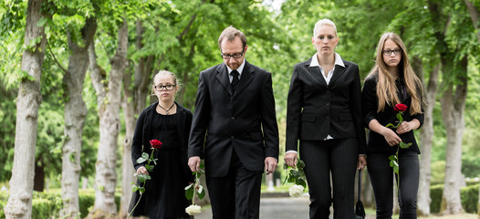 Familie läuft auf Allee im Friedhof in Trauer mit Blumen in der Hand