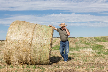 horizontal image of a farmer wearing a cowboy hat leaning against a big hay bale talking on his...