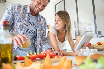 Cheerful couple in kitchen cooking dinner, using tablet