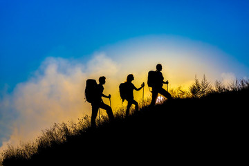 Fototapeta premium Family journey wild landscape Silhouettes of three people walking with backpacks hiking gear up toward top of wild grass mountain mother father daughter bright luminous sunrise sky background