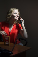 Beautiful sexy blonde woman secretary with curly hair in red blouse sitting in office at table speaking by phone and holding sand glass, vertical picture