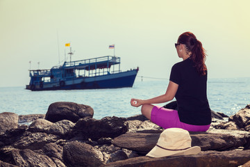 Woman sitting in lotus position, meditating, in from of ocean at sunset. Fishing boat is in background