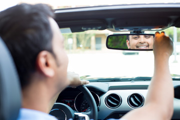 Closeup portrait, happy young man driver looking at rear view mirror smiling, adjusting image reflection, isolated interior car windshield background. Positive human expression body language