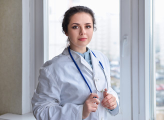 Portrait of young woman doctor on white coat standing near window