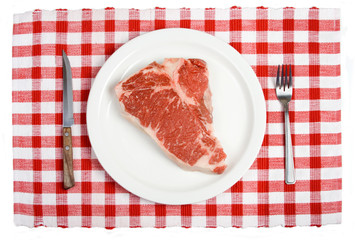 Raw T-Bone Steak on Plate – A raw T-bone steak on a white plate. With knife and fork and red checked tablecloth in background.