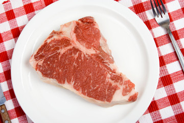 Raw T-Bone Steak – Uncooked T-bone steak on a white plate. Red checked tablecloth in background.