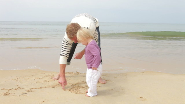 Father And Young Daughter At Beach Drawing Pictures In Sand