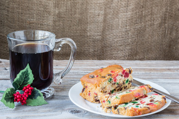 horizontal image of a freshly baked christmas fruit cake  sliced on a white plate with a clear glass cup of hot coffee adorned with a small red holly on an old rustic wood and burlap background