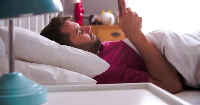 Man Waking Up In Bed And Checking Messages On Mobile Phone