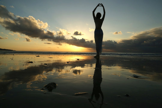 Silhouette of a young woman creating the shape of a heart with her hands against sunset background