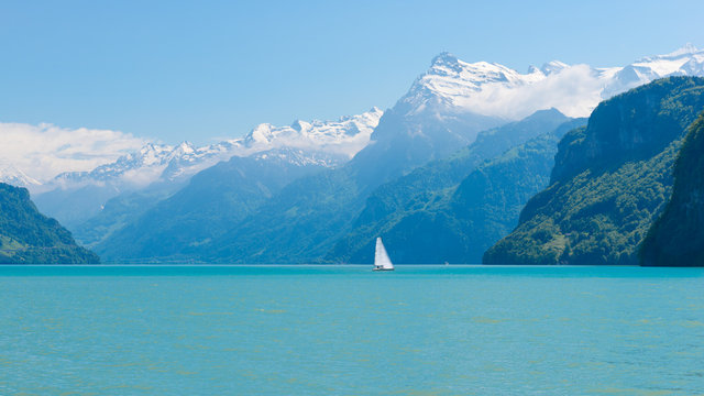Magnificent panorama of mountains with snowy peaks. On  surface of lake sailboat sails. Cloudless blue sky and a few clouds in the mountains