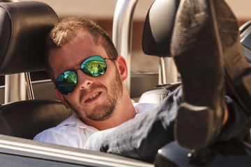Mann mit Sonnenbrille sitzt in seinem Auto mit hochgelegten Beinen