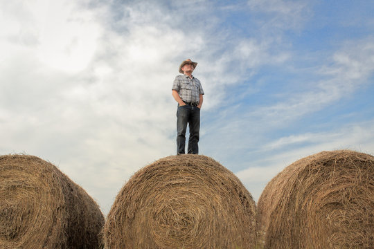 horizontal image of one farmer wearing a cowboy hat standing on a huge hay bale with his hands in his pockets gazing across the land under a beautiful cloud filled sky in the summer time