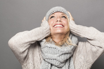 lovely young woman with blonde hair wearing winter hat and clothes, looking up with hands on ears...