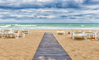 Sandy beach on cloudy day with wooden pathway and empty sun beds