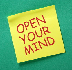 The phrase Open Your Mind in red text on a yellow sticky note posted on a green notice board as a reminder