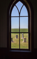 horizontal image of an inside view looking out of an arched church window at a grave yard sitting on green grass and under a clear blue sky in the summer time.