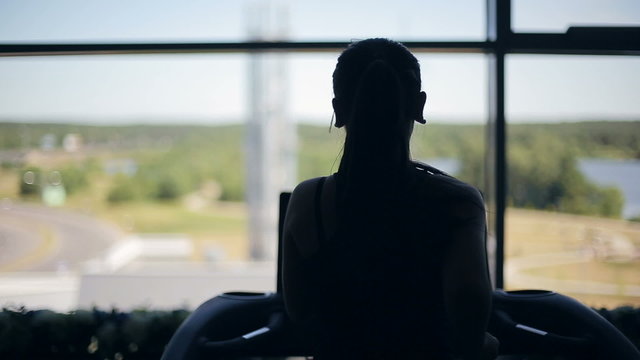 Silhouette of girl running on the treadmill and looking into the large window. Left to right dolly shot
