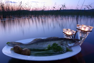 Fresh fish catch (pike perch) on plate served with fresh vegetables in outdoors setting on a lake with candles as a decoration at sunset time on summer evening at Nokia, Finland.
