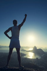 Fototapeta na wymiar Inspirational silhouette of athlete in white sport uniform standing with champion arm raised in front of Rio de Janeiro Brazil sunrise skyline overlook at Sugarloaf Mountain