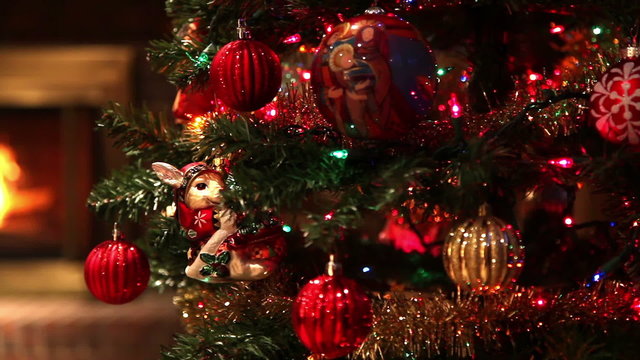 Tracking Across A Christmas Tree Full Of Ornaments Standing In A Living Room, Revealing An Out Of Focus Fire Burning In A Fireplace In The Background.