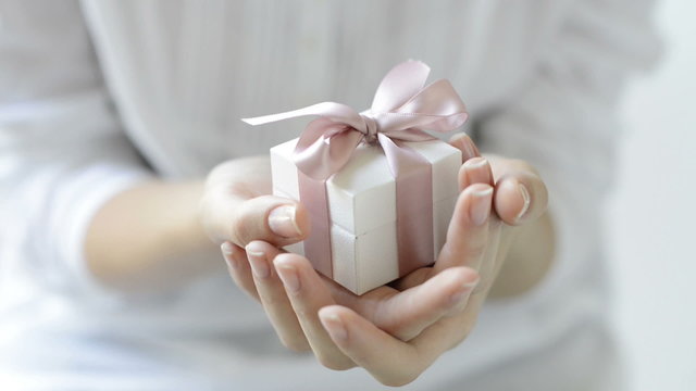 Close up shot of female hands holding a small gift wrapped with pink ribbon. Small gift in the hands of a woman indoor. Shallow depth of field with focus on the little box.