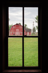 vertical image of the view from inside looking out a four pane window at a red barn with a windmill and lots of green grass in the summer time