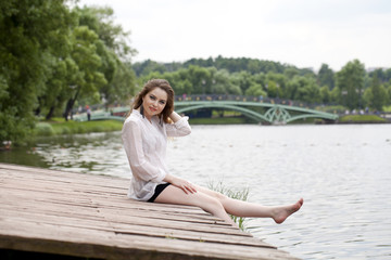 Fototapeta na wymiar Young beautiful girl in a white tunic sits on a wooden pier on t