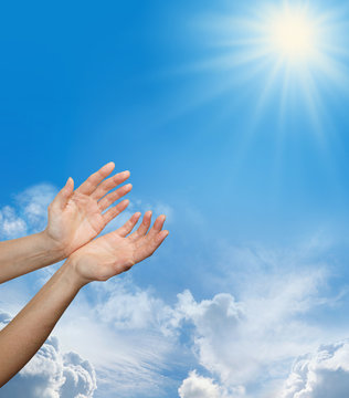 Divine Source -  Female hands reaching up towards a bright sun burst on a blue sky background with fluffy clouds and plenty of copy space