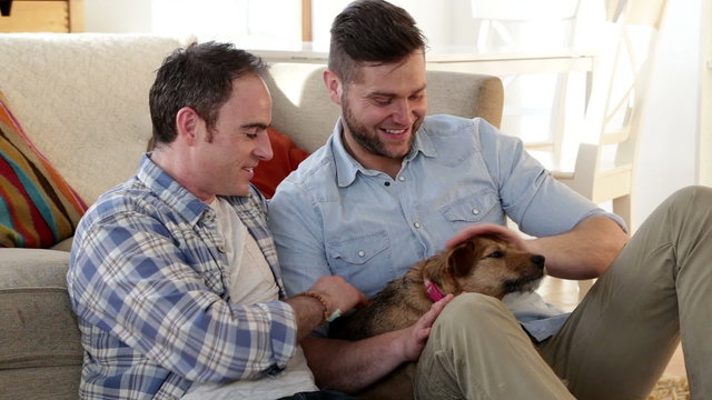 Same sex couple sitting at home together, petting their dog and talking.