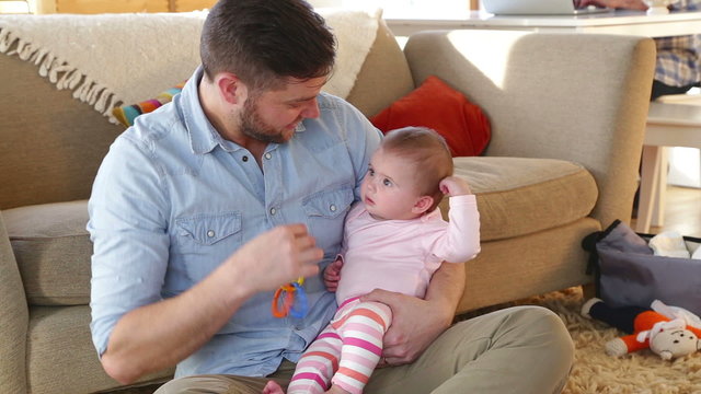 Father sitting in his home with his baby daughter. He is playing with her toys with her. 