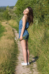Cute brunette girl in denim shorts and white sneakers standing on the road in the countryside in summer