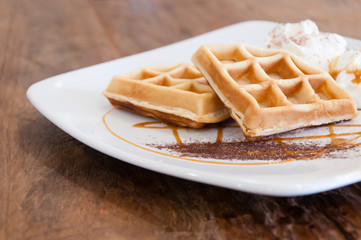 Two waffles on dish, decorated with caramel sauce and whipping cream on wood table