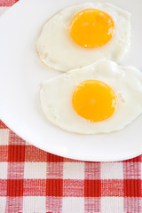 Two Fried Eggs on Plates – Two fried, sunny side up eggs on a white plate. Red checked background.