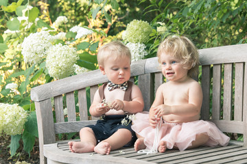 Baby boy and girl in formal dress sitting on wooden bench in a beautiful garden