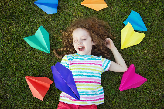 Happy Laughing Girl Throwing Paper Airplane In Green Grass At Su