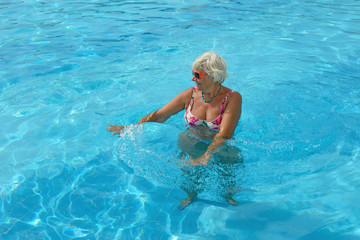 Fototapeta na wymiar Aged woman is doing spa exercises in bright blue water of pool.