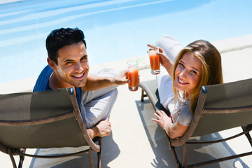 beautiful young couple having drink in deck chairs by the pool in holiday resort