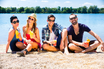 Vier Freunde sitzen mit Getränken am See Strand in der Sommersonne
