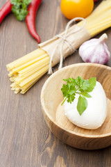 Noodles in paper tied with a rope, a wooden bowl mozzarella, fresh herbs and fresh vegetables on wooden table