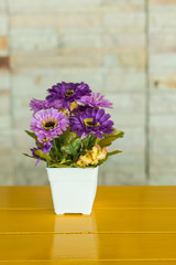 Purple artificial flowers in a white vase rests on yellow wooden