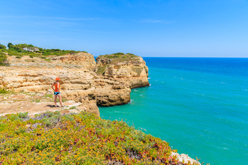 Young woman tourist standing on cliff and looking at beautiful beach near Carvoeiro town, Algarve region, Portugal