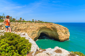 Young woman tourist standing on cliff rock and looking at sea cave on coast of Portugal, Algarve...